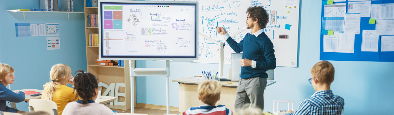 Elementary School Science Teacher Uses Interactive Digital Whiteboard to Show Classroom Full of Children how Software Programming works for Robotics. Science Class, Curious Kids Listening Attentively (Elementary School Science Teacher Uses Interactive